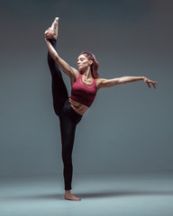 Flexible young female dancer practices stretching in stylish sportswear barefoot in studio isolated on gray background
