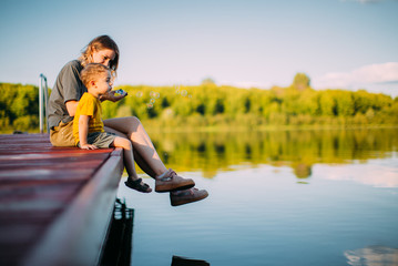 Young mother and baby boy sitting on dock launch soap bubbles. Summer photography for blog or ad...