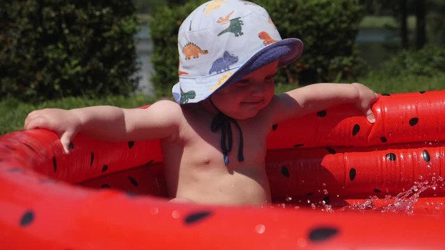 Cute baby in bucket hat relaxing in inflatable watermelon-style swimming pool. High quality FullHD footage