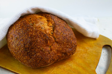 Freshly baked cereal bread on a wooden board under a white towel.