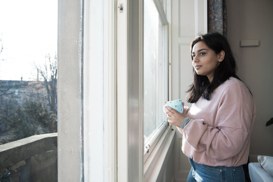 A British-Bangladeshi woman wearing jeans and a pink top enjoys a cup of tea while standing next to a window and looking out in a flat in Edinburgh, Scotland, United Kingdom