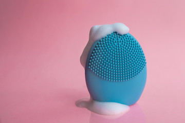 Silicone brush for washing face in foam. Blue facial cleansing brush on pink background. Beauty and skincare concept. Brush for face lifting, anti-aging wrinkles and massage