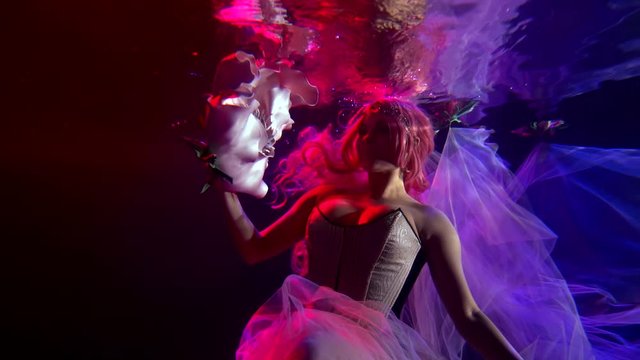 a woman with red hair in a corset floats in the dark under the water. she moves her hand beautifully with a very large flower. contoured pink and purple light. average plan