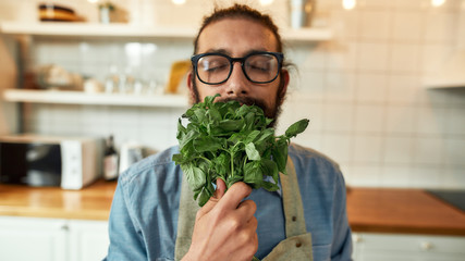 Close up of young man, Italian cook in apron smelling basil leaves while getting ready to prepare a meal, standing in the kitchen. Cooking at home concept