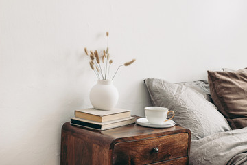 Modern white ceramic vase with dry Lagurus ovatus grass and cup of coffee on retro wooden bedside...