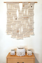 Hand crafted macrame wall decoration with wooden elements. hanging on a wall and below placed on the shelf three hand made candle holders, big size in the middle.