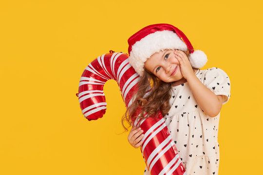 A cute little girl child in a smart dress and a Santa hat holds an inflatable shape of a candy cane on a yellow background. 2021 new year holidays concept and childrens new year gifts advertising