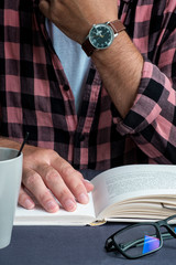 A man in a plaid shirt is reading a book at the table. Glasses, watch and cup.