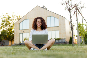 Full length frontal portrait of a smiling young woman with curly hair with laptop seated on grass in the her yard.