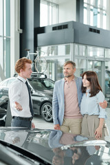 Obraz premium Confident young sales manager in formalwear showing one of new cars to clients