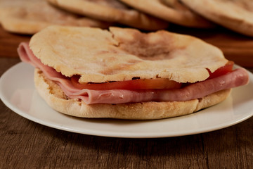 Close up of Pita bread sandwiches with ham and tomatoes. Served sandwich served on a white earthenware plate and on a wooden board there are more breads.
