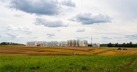 Panorama of large wheat field of cereals during the harvest with combine, people and tractor, against backdrop of new urban neighborhood with crane while building house with blue sky and clouds.