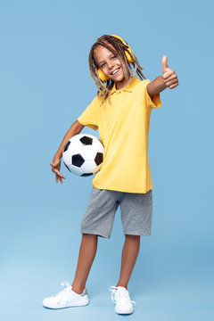Full lengh image of cute little boy with african dreads holding soccer ball and show thumb up over blue background.