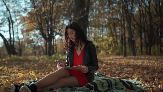 attractive curly brunette woman sitting and writing in notepad in park at fall
