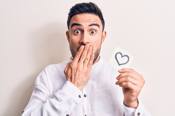 Young handsome romantic man with beard holding reminder with heart shape symbol covering mouth with hand, shocked and afraid for mistake. Surprised expression