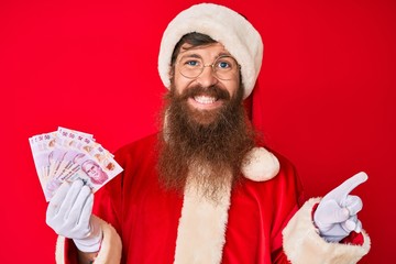 Handsome young red head man with long beard wearing santa claus costume holding mexican pesos smiling happy pointing with hand and finger to the side
