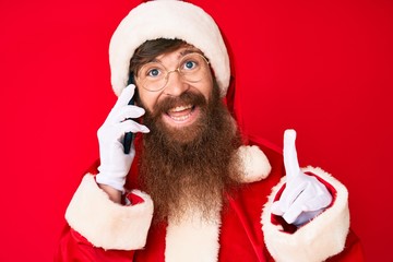 Handsome young red head man with long beard wearing santa claus costume talking on the phone surprised with an idea or question pointing finger with happy face, number one