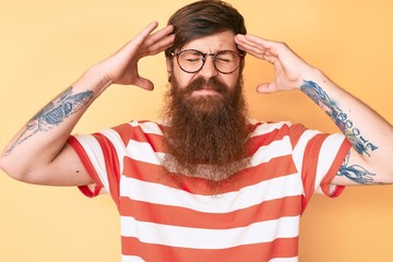 Handsome young red head man with long beard wearing casual clothes and glasses with hand on head,...