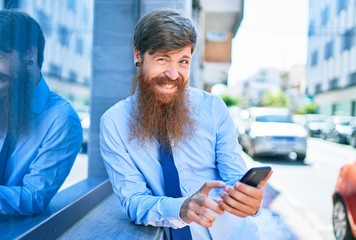 Young handsome redhead businessman wearing elegant clothes smiling happy. Leaning on the wall with smile on face using smartphone at street of city.