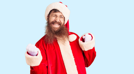 Handsome young red head man with long beard wearing santa claus costume very happy and excited doing winner gesture with arms raised, smiling and screaming for success. celebration concept.