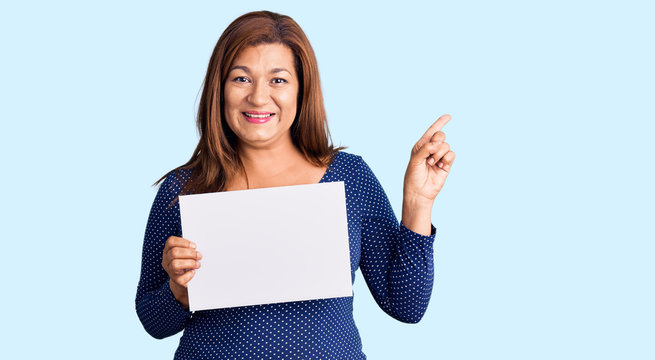 Middle age latin woman holding paper banner with blank space smiling happy pointing with hand and finger to the side
