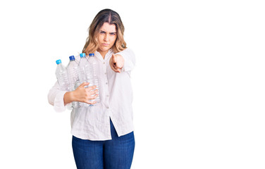 Young caucasian woman holding recycling plastic bottles pointing with finger to the camera and to you, confident gesture looking serious