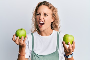 Beautiful caucasian woman holding green apples angry and mad screaming frustrated and furious, shouting with anger. rage and aggressive concept.