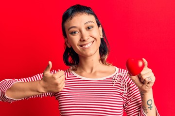 Young woman holding heart smiling happy and positive, thumb up doing excellent and approval sign