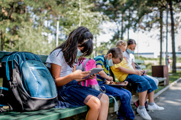 Back to school during quarantine. Children with backpacks wearing protective face mask and using mobile phones and digital tablets.