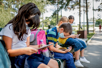 Fototapeta na wymiar Back to school during quarantine. Children with backpacks wearing protective face mask and using mobile phones and digital tablets.