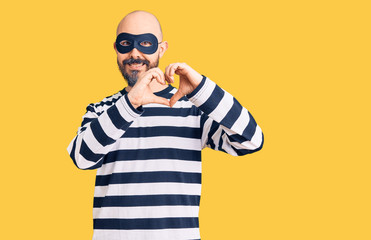 Young handsome man wearing burglar mask smiling in love doing heart symbol shape with hands. romantic concept.