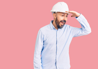 Young handsome man wearing architect hardhat very happy and smiling looking far away with hand over head. searching concept.