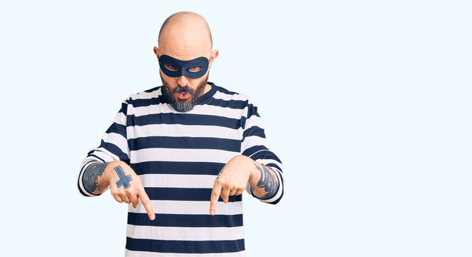 Young handsome man wearing burglar mask pointing down with fingers showing advertisement, surprised face and open mouth