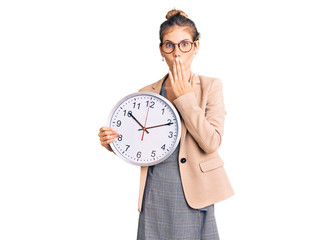 Beautiful caucasian woman with blonde hair wearing business jacket and holding clock covering mouth...