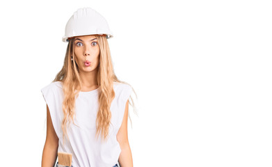 Fototapeta premium Beautiful caucasian woman with blonde hair wearing hardhat and painter clothes scared and amazed with open mouth for surprise, disbelief face