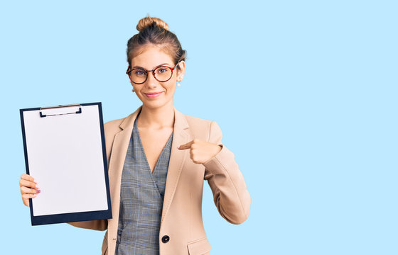 Beautiful caucasian woman with blonde hair holding clipboard with blank space pointing finger to one self smiling happy and proud