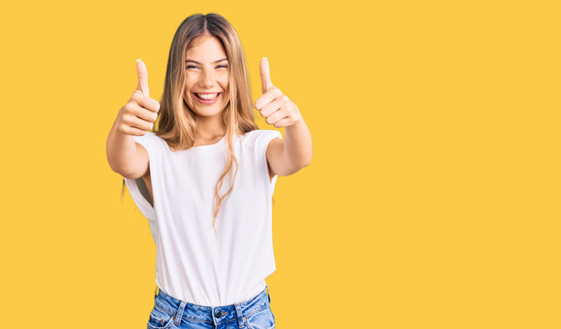 Beautiful caucasian woman with blonde hair wearing casual white tshirt approving doing positive gesture with hand, thumbs up smiling and happy for success. winner gesture.