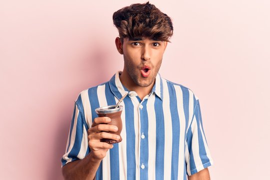 Young hispanic man drinking mate infusion scared and amazed with open mouth for surprise, disbelief face