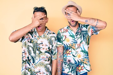 Young gay couple of two men wearing summer hat and hawaiian shirt peeking in shock covering face and eyes with hand, looking through fingers with embarrassed expression.