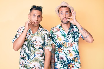 Young gay couple of two men wearing summer hat and hawaiian shirt mouth and lips shut as zip with...