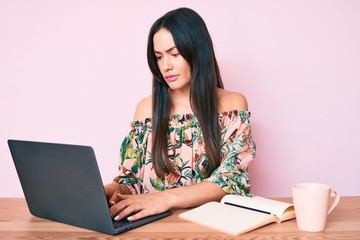 Young caucasian woman sitting at the desk studying using laptop and book drinking coffee thinking attitude and sober expression looking self confident