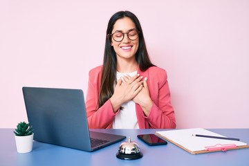Young caucasian woman sitting at the recepcionist desk working using laptop smiling with hands on chest with closed eyes and grateful gesture on face. health concept.