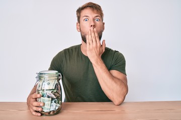Young caucasian man holding jar with savings sitting on the table covering mouth with hand, shocked...