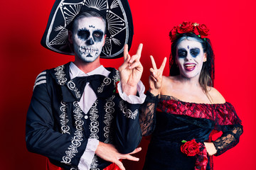 Young couple wearing mexican day of the dead costume over red smiling with happy face winking at the camera doing victory sign. number two.