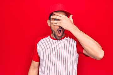 Young handsome player man wearing baseball sportswear over isolated red background peeking in shock covering face and eyes with hand, looking through fingers afraid