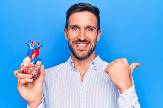 Young handsome man holding heart organ with veins and arteries over blule background pointing thumb up to the side smiling happy with open mouth
