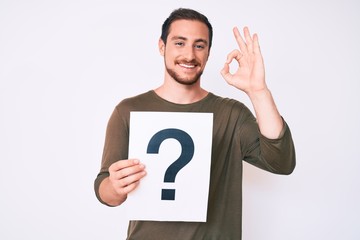 Young handsome man holding question mark doing ok sign with fingers, smiling friendly gesturing excellent symbol