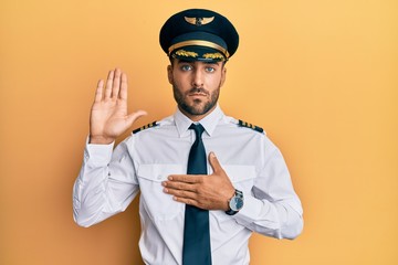 Handsome hispanic man wearing airplane pilot uniform swearing with hand on chest and open palm,...
