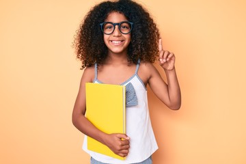 African american child with curly hair wearing glasses and holding book smiling with an idea or question pointing finger with happy face, number one