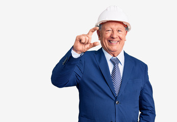 Senior handsome grey-haired man wearing suit and architect hardhat smiling and confident gesturing with hand doing small size sign with fingers looking and the camera. measure concept.